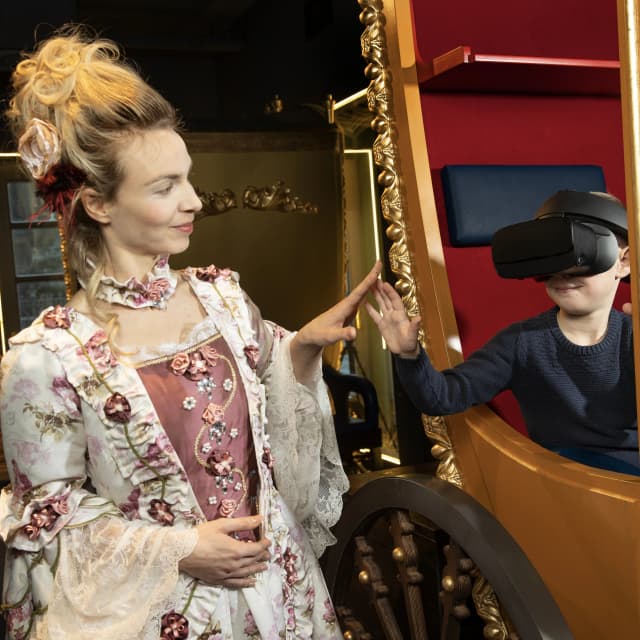 Historisch gekleidete Frau interagiert mit einem Kind, das in einer Kutsche sitzt und eine VR-Brille trägt, im TimeRide Dresden
